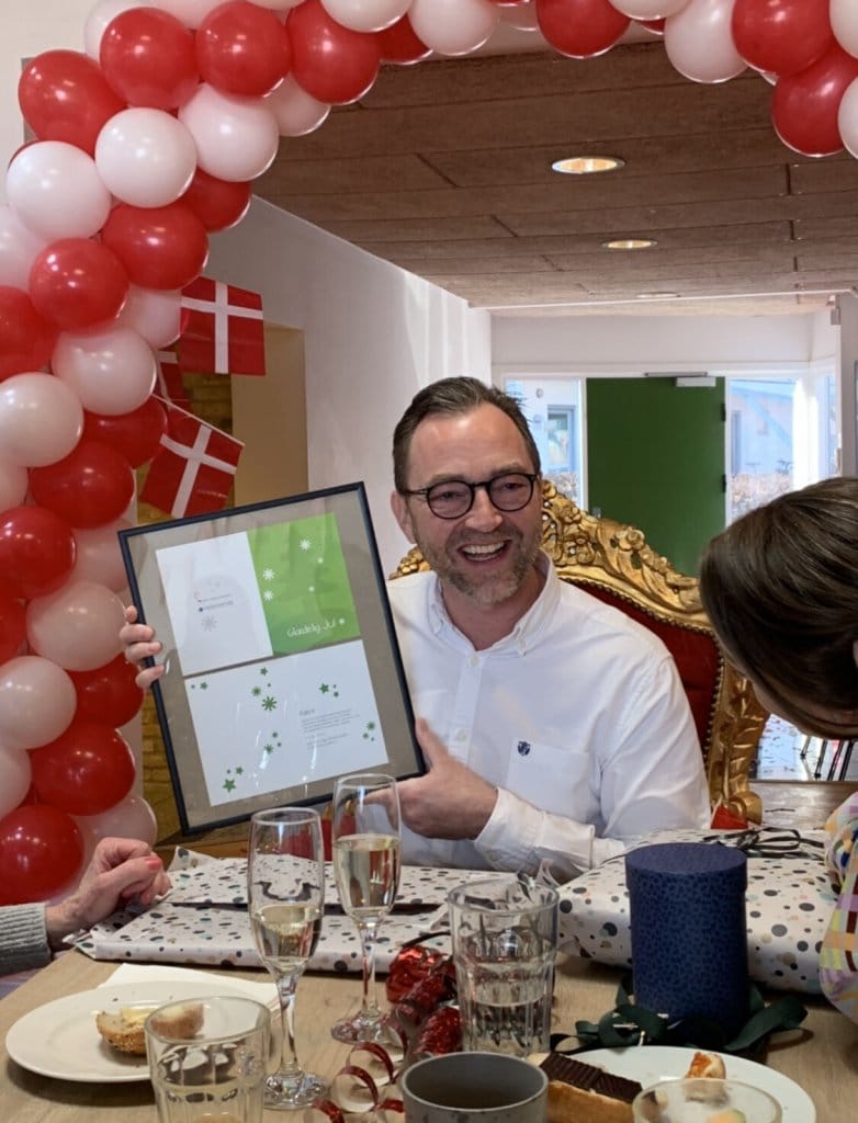 Soren Arnsbo holding a framed certificate at a celebration table with Danish flags and a balloon arch